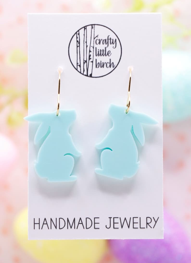 K&ouml;nnte beinhalten: Ein Paar hellblaue Ohrringe in Hasenform mit goldenen Kreisen. Die Ohrringe befinden sich auf einer wei&szlig;en Karte mit dem Text "crafty little birch" und "HANDMADE JEWELRY".