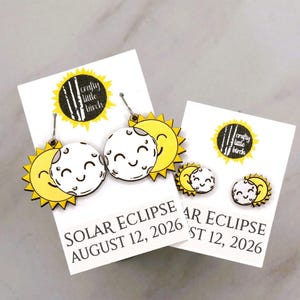 May include: Two pairs of earrings featuring a yellow sun and white moon with smiling faces in a solar eclipse design. The display cards are printed with the text SOLAR ECLIPSE AUGUST 12, 2026 and the logo for Crafty Little Birch in black and yellow.