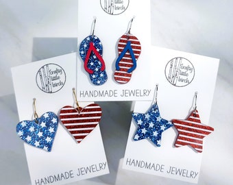 Op de afbeelding: Vier paar handgemaakte oorbellen met rood, wit en blauw glitterontwerpen. De ontwerpen omvatten slippers, harten en sterren. Elk paar is gemarkeerd met "Handgemaakte sieraden" en "Crafty Little Birch".