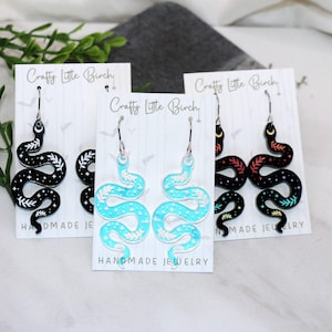 May include: Three pairs of dangle earrings featuring a snake design. The earrings are made of black, blue, and red acrylic. The snake designs are outlined in white and have a starry sky pattern. The earrings are labeled "Crafty Little Birch" and "Handmade Jewelry".