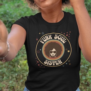 May include: A black t-shirt featuring a retro-style graphic of a woman with an afro wearing sunglasses. The graphic is surrounded by a circle with the text "Funk Soul Sister" in a rainbow font.