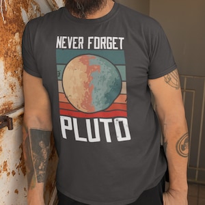 May include: A dark gray t-shirt with a retro-style graphic of Pluto. The graphic features a stylized image of Pluto with a red, white, and blue color scheme. The text "Never Forget Pluto" is printed in white above the graphic.