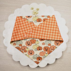 May include: A Halloween-themed pet bandana featuring a pattern of smiling pumpkins in orange, yellow, and gray on a cream background. The top section is orange and tan checkered fabric, secured with two pink buttons.