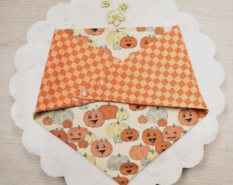 Peut inclure: Un bandana pour animaux de compagnie sur le thème d'Halloween, avec un motif de citrouilles souriantes orange, jaune et grise sur fond crème. La partie supérieure est en tissu à carreaux orange et beige, fixée par deux boutons roses.