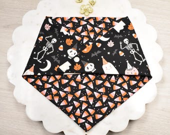 Puede incluir: Un pañuelo para mascotas con temática de Halloween con fondo negro. La mitad superior presenta esqueletos, fantasmas, murciélagos y caramelos de maíz. La mitad inferior tiene un patrón de caramelos de maíz. El pañuelo está doblado en forma de triángulo.