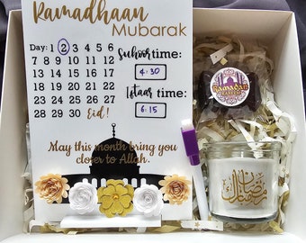 May include: A white gift box with a white card that says "Ramadhaan Mubarak" and has a calendar with the days of Ramadan. The box also contains a candle with Arabic writing, a small bag of dates, and a white dry-erase marker.