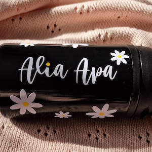 Op de afbeelding: Zwarte reistumbler met witte madeliefjesontwerpen en de naam "Alia Ava" gedrukt op de zijkant.