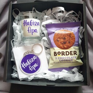 May include: A gift box filled with white tissue paper containing a clear glass candle with a purple label that says "Habiza Apa", a purple keychain with the same text, and a purple and white package of two chocolate cookies.