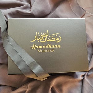 Pode incluir: Uma caixa de presente preta com uma fita dourada e o texto "Ramadhaan Mubarak" em árabe e inglês.
