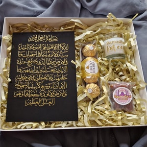 May include: A white gift box filled with gold tissue paper. The box contains a black card with gold Arabic calligraphy, a small glass candle with the words "Ramadhan Kareem" in gold lettering, a package of chocolate sweets, and a small brown box with the words "Ramadan Kareem" in gold lettering.