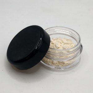 Pode incluir: Um pequeno frasco de plástico transparente com tampa preta, cheio de pó branco. A tampa está aberta e o frasco está sobre uma superfície branca.