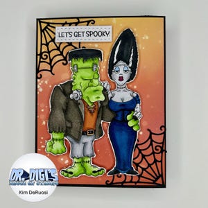 May include: A Halloween-themed greeting card featuring a cartoon illustration of Frankenstein and his bride. The card has a black border with spiderwebs, and the text "LET'S GET SPOOKY" is at the top. The background is orange and yellow.