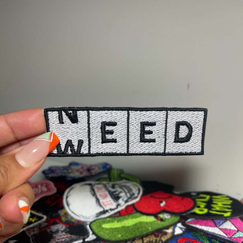 May include: Embroidered patch with the word "NEED" in white letters on a black and white background. The patch is rectangular and has a black border. Other patches are visible in the background.