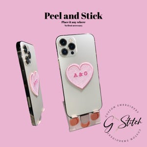 May include: Two white smartphones with a pink heart-shaped patch embroidered with "A&G". The phones are displayed on a pink background with the text "Peel and Stick" and "Place it any where No heat necessary". The product is from "G Stitch Custom Embroidery".