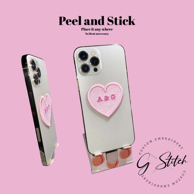 May include: Two white smartphones with a pink heart-shaped patch embroidered with "A&G". The phones are displayed on a pink background with the text "Peel and Stick" and "Place it any where No heat necessary". The product is from "G Stitch Custom Embroidery".