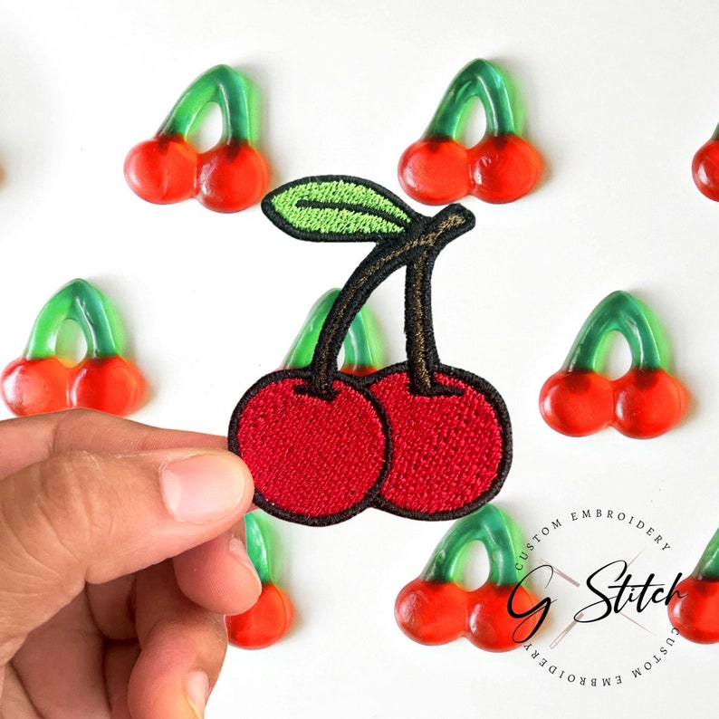 May include: A red embroidered cherry patch with a black stem and green leaf is held in a hand. The patch has a textured appearance. Scattered around the patch are gummy cherry candies with red cherries and green stems.