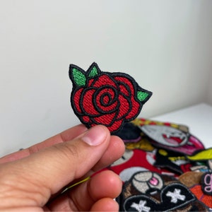May include: Embroidered patch featuring a red rose with green leaves and black outlining. The rose is in full bloom, with detailed stitching. The patch is likely for clothing or accessories.