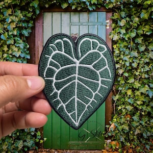 May include: A dark green, heart-shaped embroidered patch featuring a detailed white leaf vein design. The patch has a black border and the text "Custom Embroideries G Stitches" is visible at the bottom. The patch is held in front of a green door.