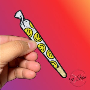 May include: Embroidered patch in the shape of a rolled cigarette. The white patch features yellow smiley faces and a tan tip. The patch is held by a hand against a gradient background of red, orange, and purple.