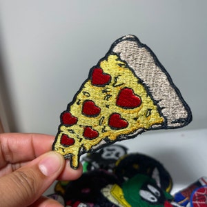 May include: Embroidered pizza slice patch with yellow cheese, red heart-shaped pepperoni, and a brown crust. The patch has a black border and is likely for clothing or accessories.