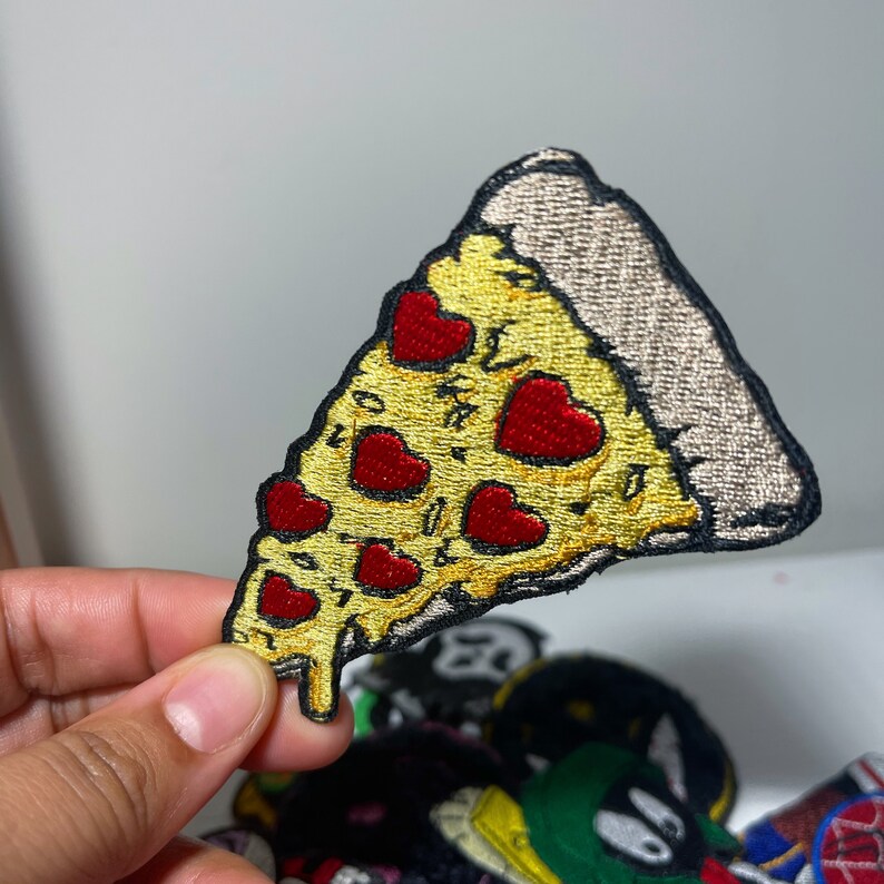 May include: Embroidered pizza slice patch with yellow cheese, red heart-shaped pepperoni, and a brown crust. The patch has a black border and is likely for clothing or accessories.