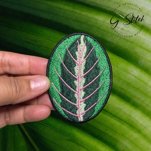 May include: Embroidered patch depicting a green leaf with pink and white veins, framed by a green oval. The patch has a black border and the text "G Stitch Custom Embroidery" is visible. The patch is held against a green leaf background.