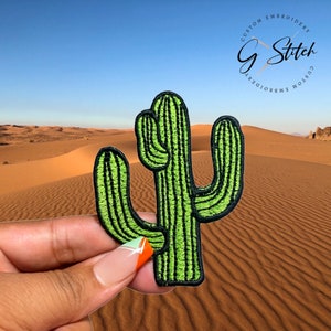 Puede incluir: Un parche de cactus verde con costuras negras, frente a un paisaje des&eacute;rtico. El parche presenta el texto "Custom Embroidery G Stitch Custom Embroidery" en un dise&ntilde;o circular.