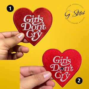 May include: Two red heart-shaped embroidered patches with the text "Girls Don't Cry" in white script. The patches are held against a yellow background. The patches are custom embroidery.