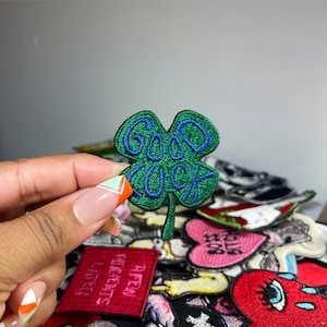 May include: Embroidered green four-leaf clover patch with the words "GOOD LUCK" in blue script. The patch is held in front of a collection of other patches with various designs and colors.