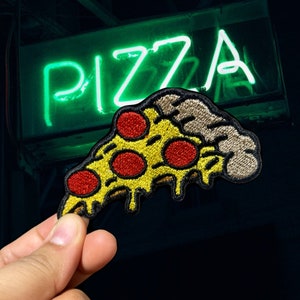May include: Embroidered pizza slice patch with yellow crust, red pepperoni, and melted cheese. The patch is held in front of a neon green "PIZZA" sign. The patch is a fun accessory for jackets, bags, or other fabric items.