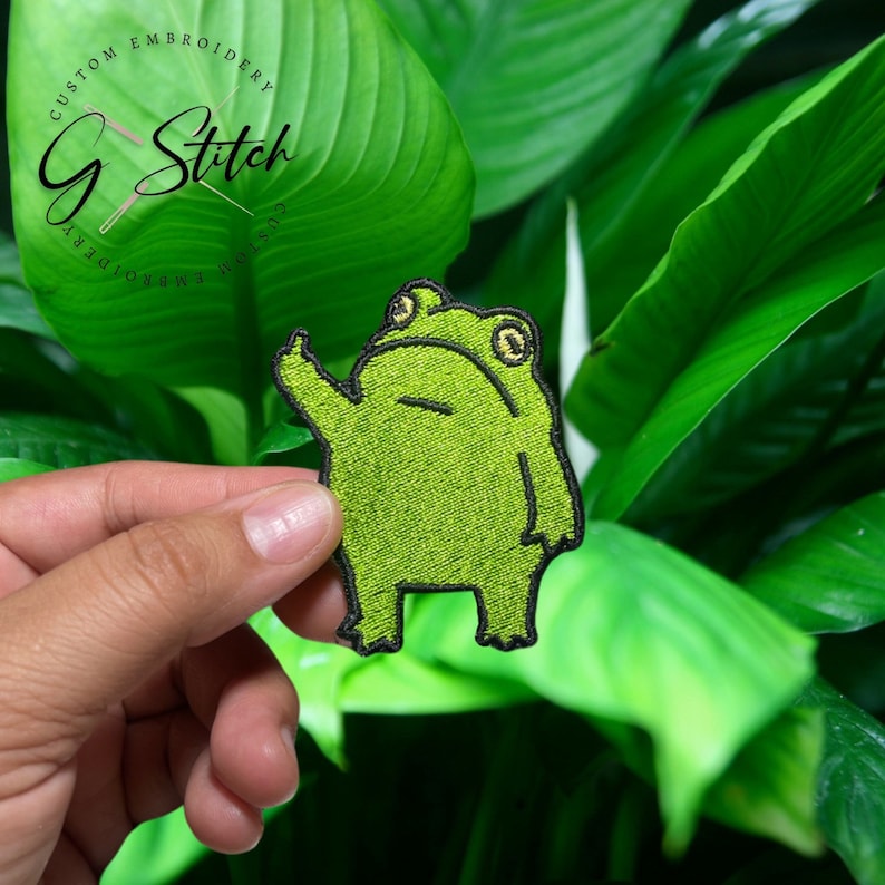 K&ouml;nnte beinhalten: Gestickter gr&uuml;ner Frosch-Aufn&auml;her mit schwarzer Umrandung, der den Mittelfinger zeigt. Der Aufn&auml;her hat gelbe Augen und befindet sich vor einem Hintergrund aus gr&uuml;nen Bl&auml;ttern. Der Text "G Stitch Custom Embroidery" steht oben.