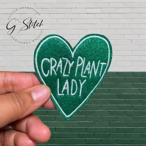 May include: A green, heart-shaped embroidered patch with white stitching and the text "CRAZY PLANT LADY". The patch is held up against a white and green brick wall. The patch is a custom embroidery item.