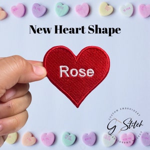 May include: A red heart-shaped embroidered patch with the word "Rose" in white. The image also features the text "New Heart Shape" and several pastel-coloured heart-shaped candies with various phrases.