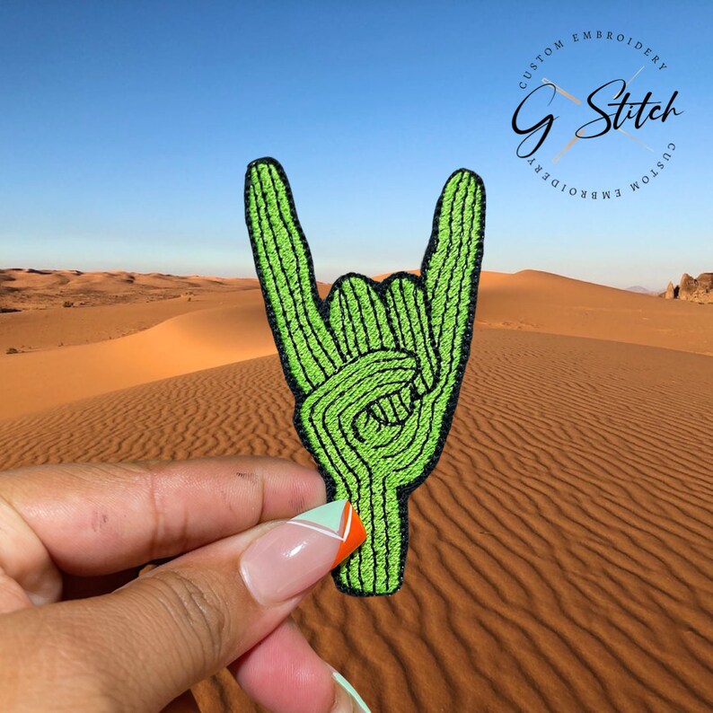 Puede incluir: Un parche bordado verde con la forma de una mano haciendo el gesto de rock and roll. El parche tiene un contorno negro y se sostiene frente a un paisaje des&eacute;rtico. El parche tiene el texto "G Stitch Custom Embroidery".