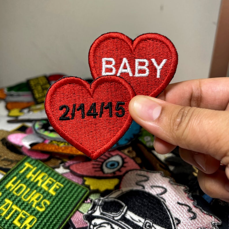 May include: Two red heart-shaped embroidered patches. One patch has the word "BABY" in white. The other patch has the date "2/14/15" in black. The patches are held in front of a collection of other patches.