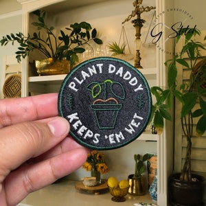May include: A circular, black embroidered patch with the words "PLANT DADDY KEEPS 'EM WET" in white. It features a green and orange plant in a pot. A humorous accessory for plant enthusiasts.