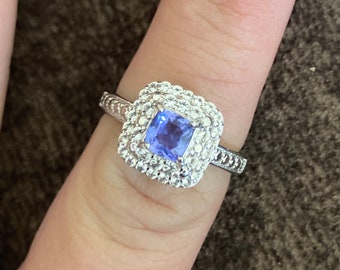Op de afbeelding: Een zilveren ring met een vierkante blauwe edelsteen omgeven door een dubbele halo van kleinere heldere edelstenen.