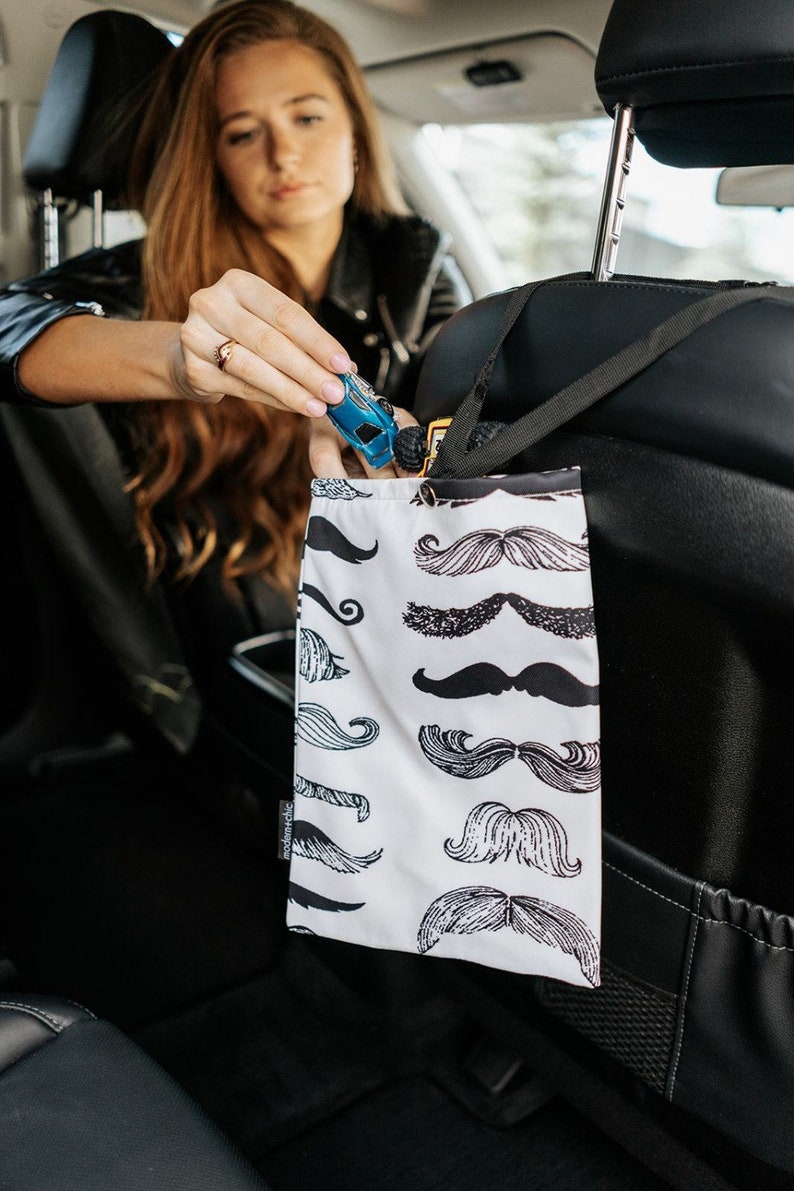 May include: A white car seat organiser with a black strap and a black and white pattern of moustaches. The organiser is hanging on the back of a car seat.
