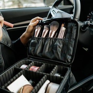 May include: Black makeup case with a zipper closure, open to reveal a variety of makeup brushes and makeup products. The case has a compartment for brushes and a separate compartment for makeup products.