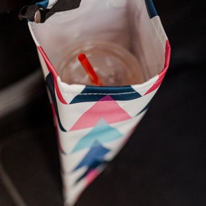 May include: A white, pink and blue geometric patterned cup holder with a black strap. A red straw is in a clear cup inside the holder.