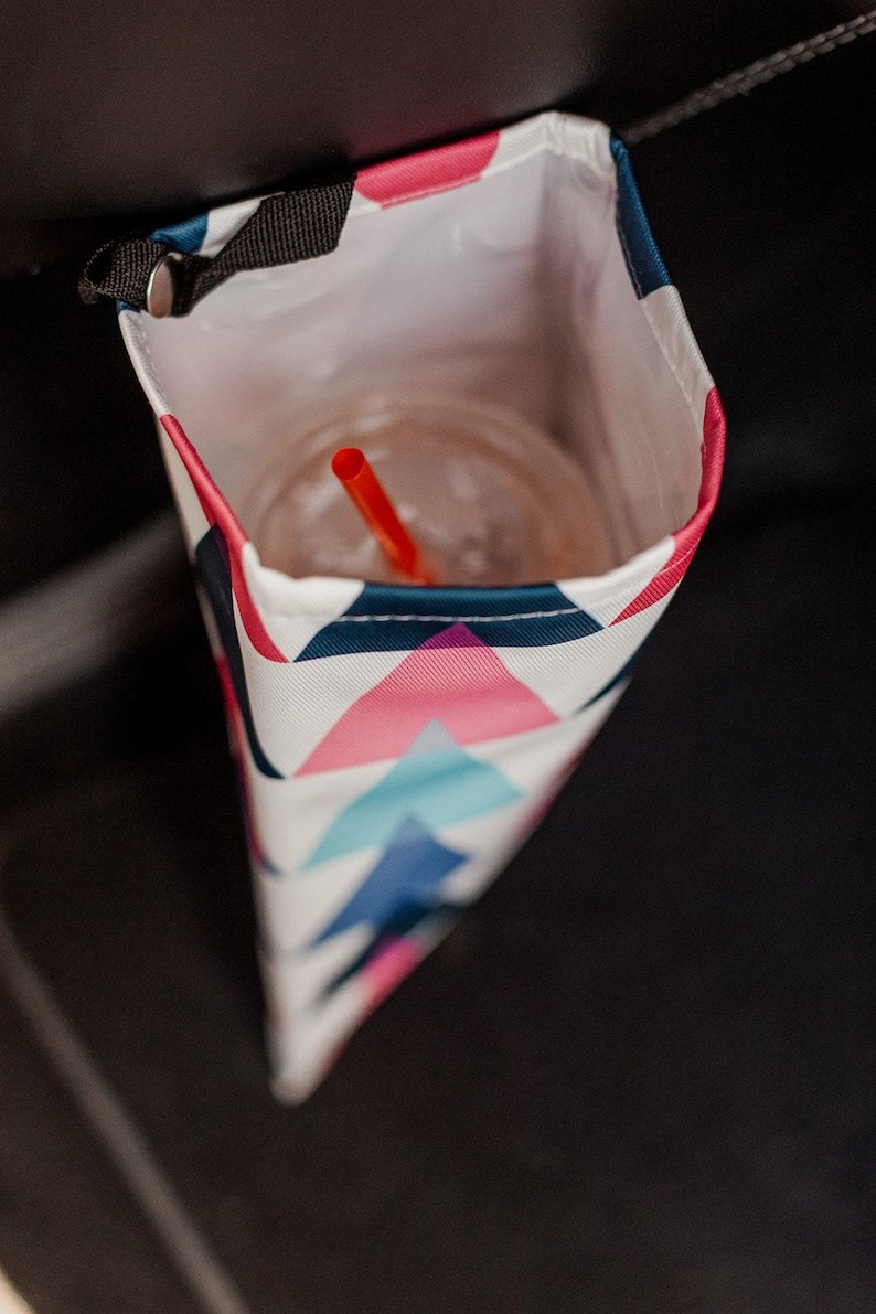 May include: A white, pink and blue geometric patterned cup holder with a black strap. A red straw is in a clear cup inside the holder.