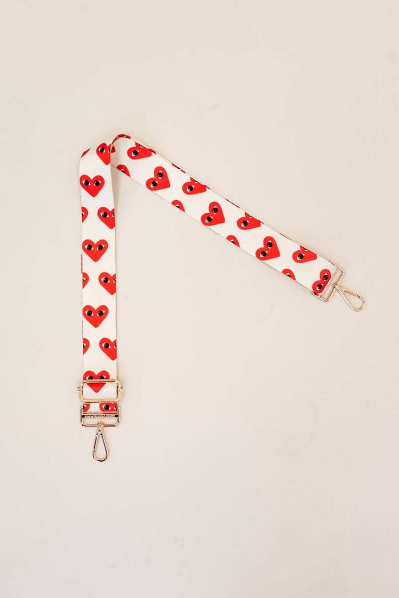 A white bag strap with red hearts with eyes and gold hardware.