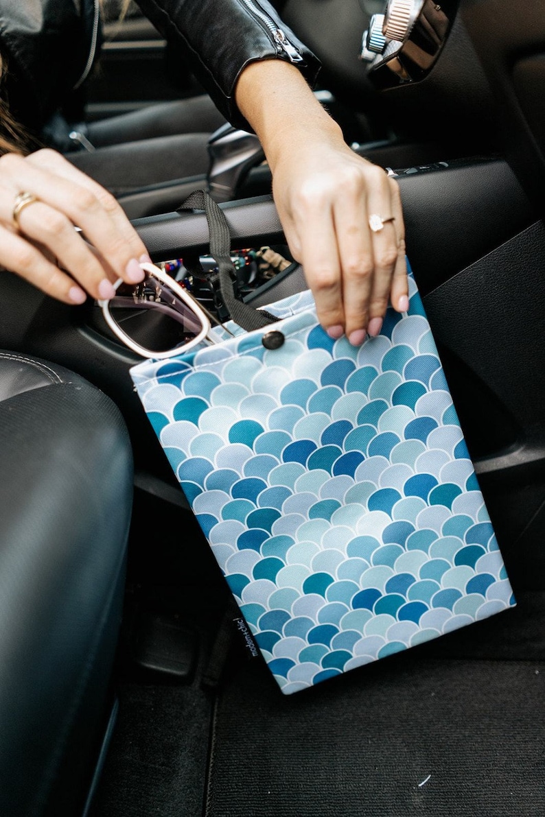 May include: A blue and white patterned car trash bag with a snap closure. The bag is hanging from a headrest in a car.