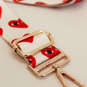 A white bag strap with red hearts with eyes and gold hardware. The gold hardware says modern+chic.