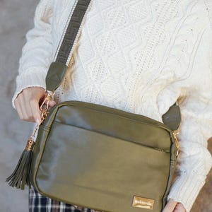 May include: Olive green crossbody bag with a matching adjustable strap and gold-tone hardware. The bag features a front pocket, a tassel, and a small gold plaque that reads "modern+chic". The bag is held by a person wearing a white sweater.