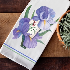 May include: White kitchen towel featuring a watercolor illustration of purple iris flowers with green leaves. The towel has blue and green stripes at the bottom and is folded on a wooden cutting board.