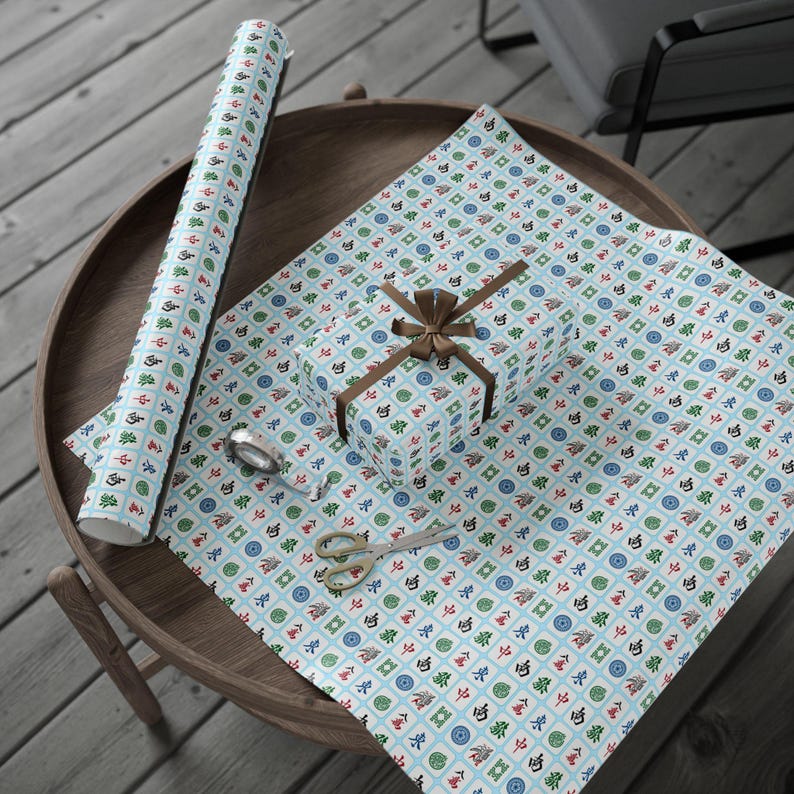 Wrapped package, and wrapping paper. the paper has white Mahjong tiles on a light blue background. The package has a string bow. Also on the table are a pair of scissors and greenery.