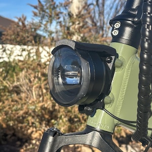 May include: Close-up of an electric bicycle headlight. The round black headlight is mounted on a green frame. The image is taken outdoors with a blurred background of trees and houses.
