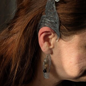 May include: A clear, gray ear cuff and earring set. The ear cuff is shaped like a leaf with intricate details, and the earring features a clear, leaf-shaped pendant with a chain and a cluster of beads in various colors.