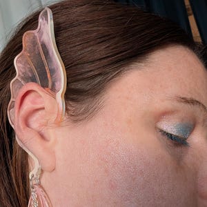 May include: A clear, pink ear cuff with a wing-like design is worn on the ear. The cuff has a dangling earring with beads. The person has brown hair and is wearing eye makeup.